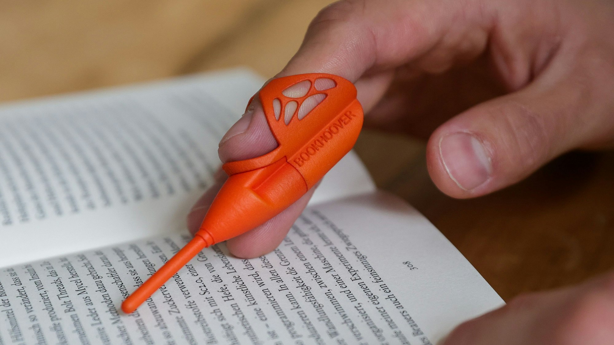 Das Foto zeigt ein aufgeschlagenes Buch und zwei Hände in Nahaufnahme. Die fotografierte Person hat auf ihren Finger das Lese-Werkzeug „Bookhoover“ aufgesteckt.