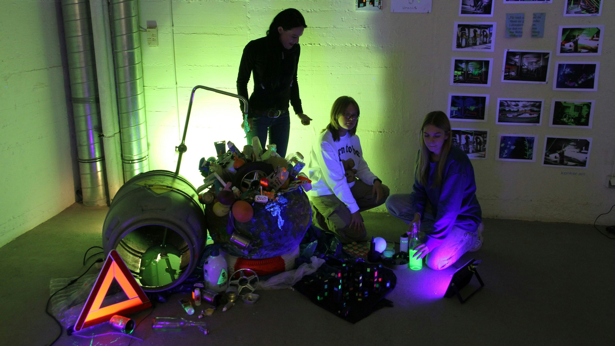 Eine Frau und zwei jugendliche Schülerinnen vor einer grün erleuchteten Plastik aus verschiedenen Müllteilen