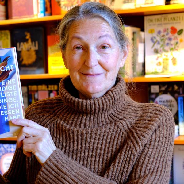Iris Pütz steht vor einem Buchregal. Sie hat ein Exemplar eines Buchs von Doris Knecht in der Hand.