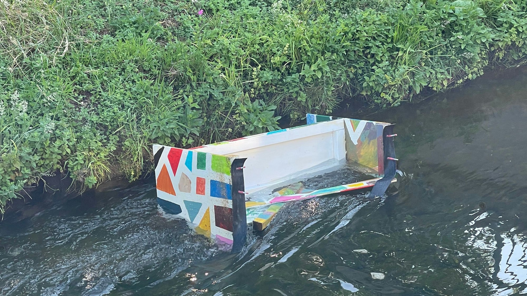 Das Bild zeigt die bunte Bank im Wasser des Veybachs.