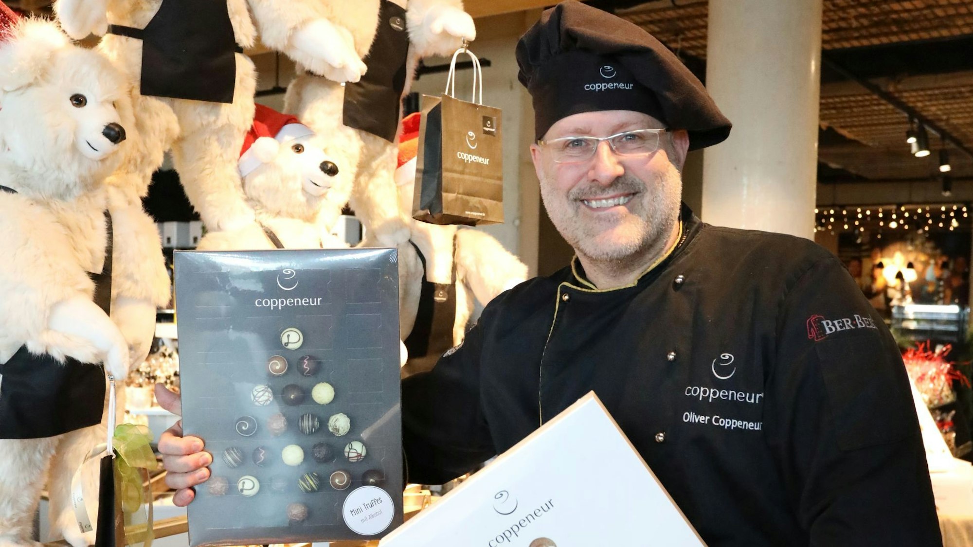 Das Bild zeigt den Gründer der Coppeneur et Compagnon, Oliver Coppeneur. Der 55-Jährige mit schwarzer Mütze hält zwei große Pralinenschachteln.