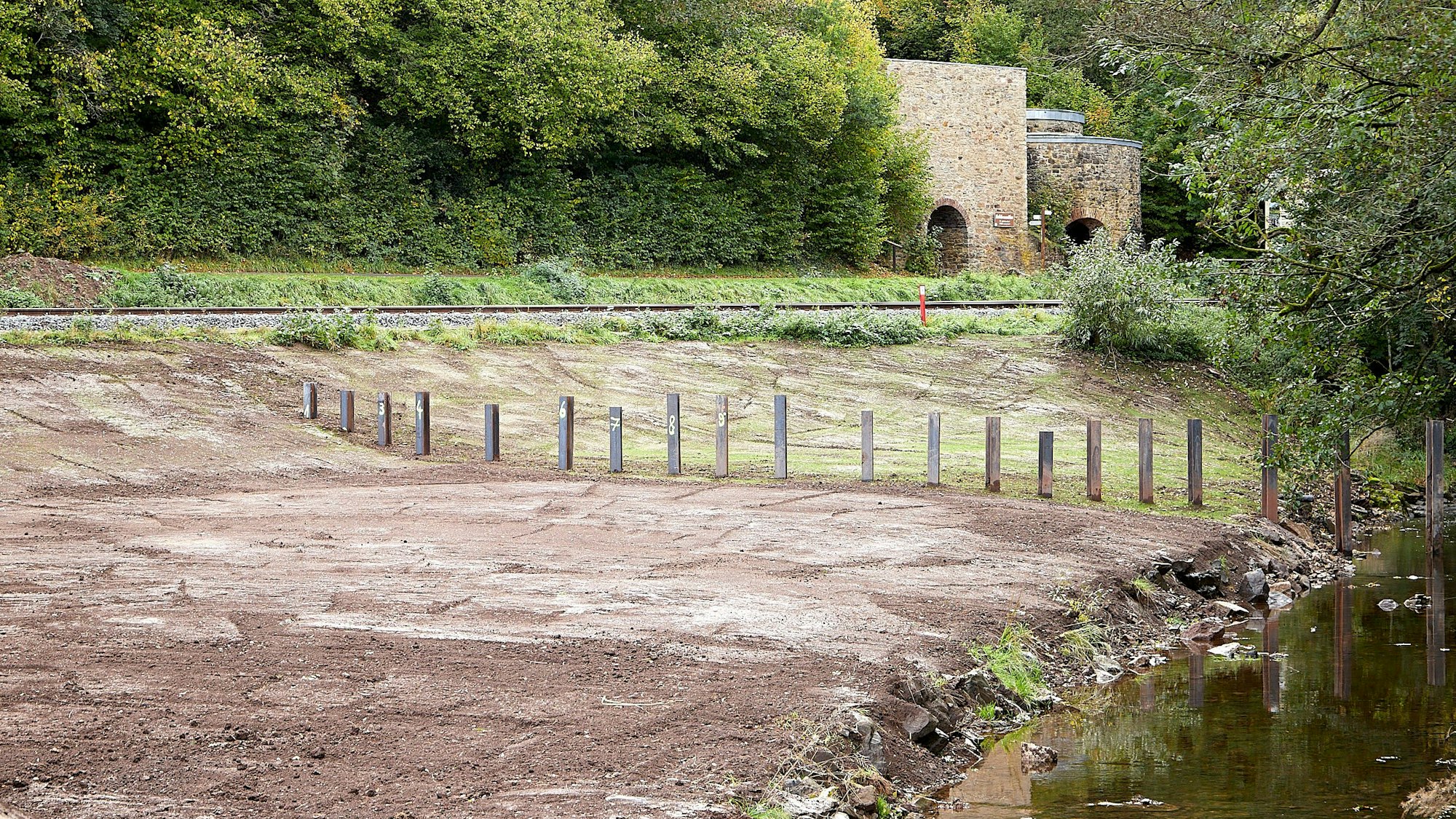 Neben dem Gewässer wurden in geringem Abstand Stahlträger vertikal in den Boden gesetzt, so dass eine Art Rechen entstanden ist.