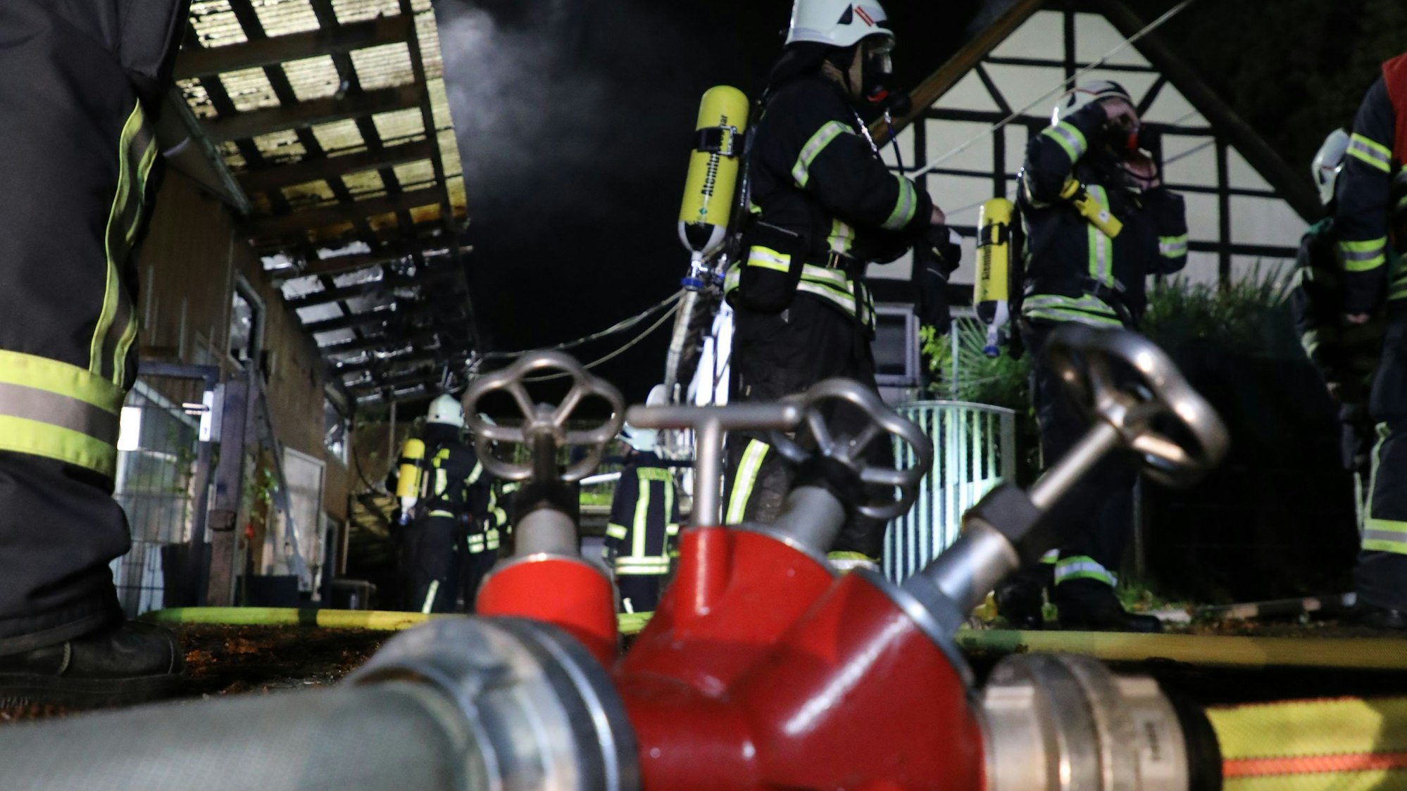 Ein Verteilerstück mit Feuerwehrschläuchen liegt auf dem Boden. Im Hintergrund sind Feuerwehrleute mit Atemschutzausrüstung zu sehen.