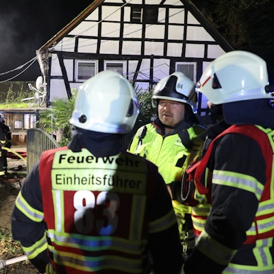 Feuerwehrleute stehen vor einem Fachwerkhaus, weitere Einsatzkräfte löschen im Hintergrund ein Nebengebäude.