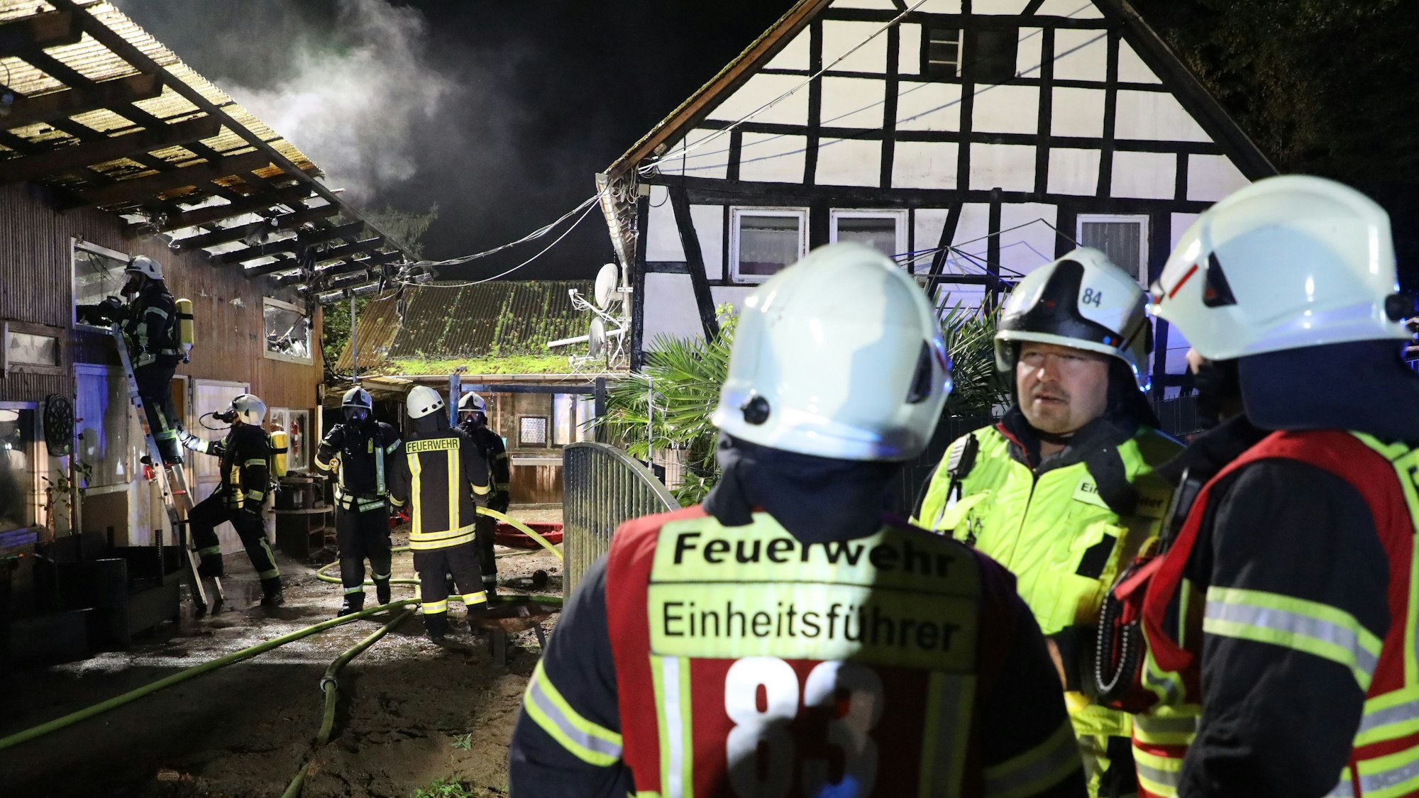 Feuerwehrleute stehen vor einem Fachwerkhaus, weitere Einsatzkräfte löschen im Hintergrund ein Nebengebäude.