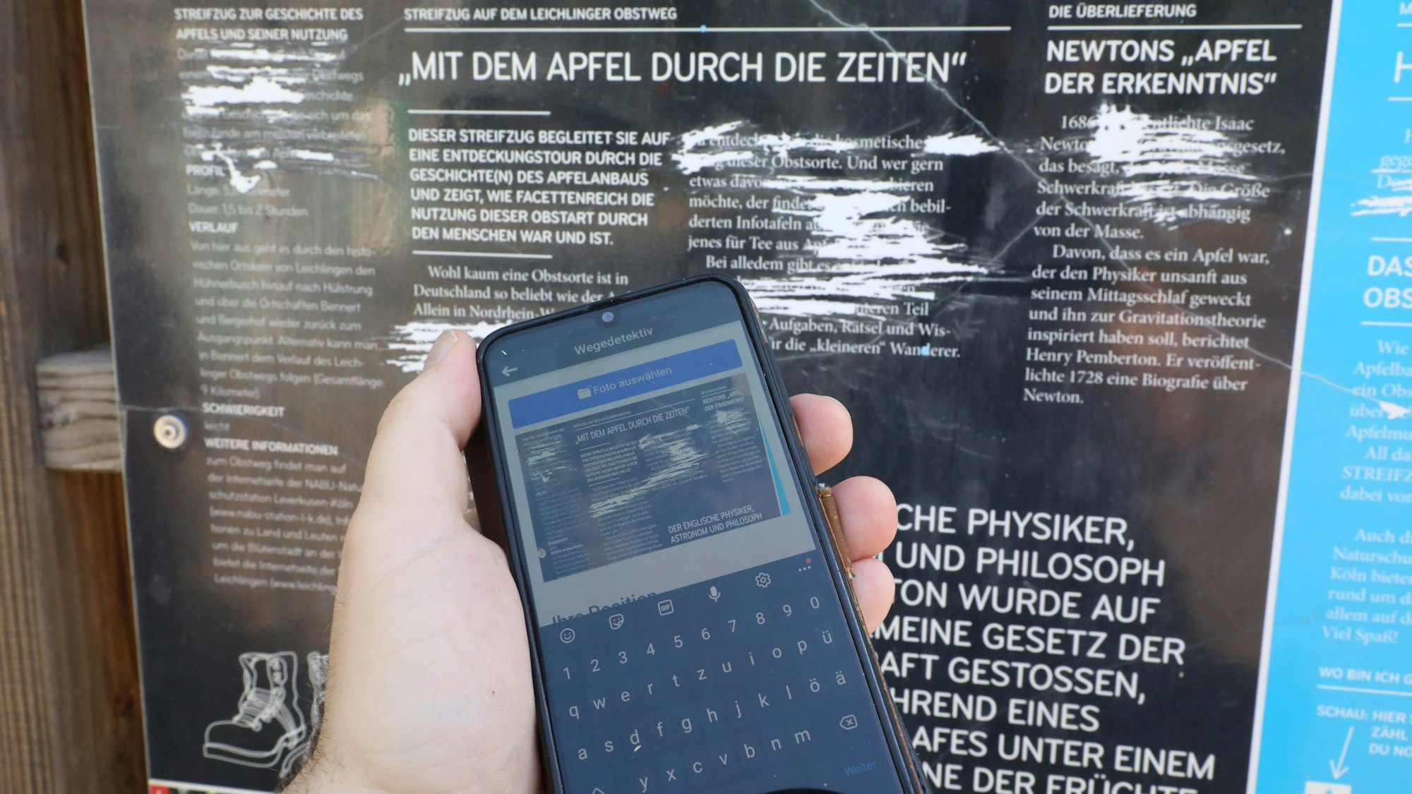 Vor einer zerkratzten Infotafel eines Bergischen Streifzug ist ein Handy zu sehen, mit dem gerade ein Foto für eine Meldung des Schadens aufgenommen wurde.