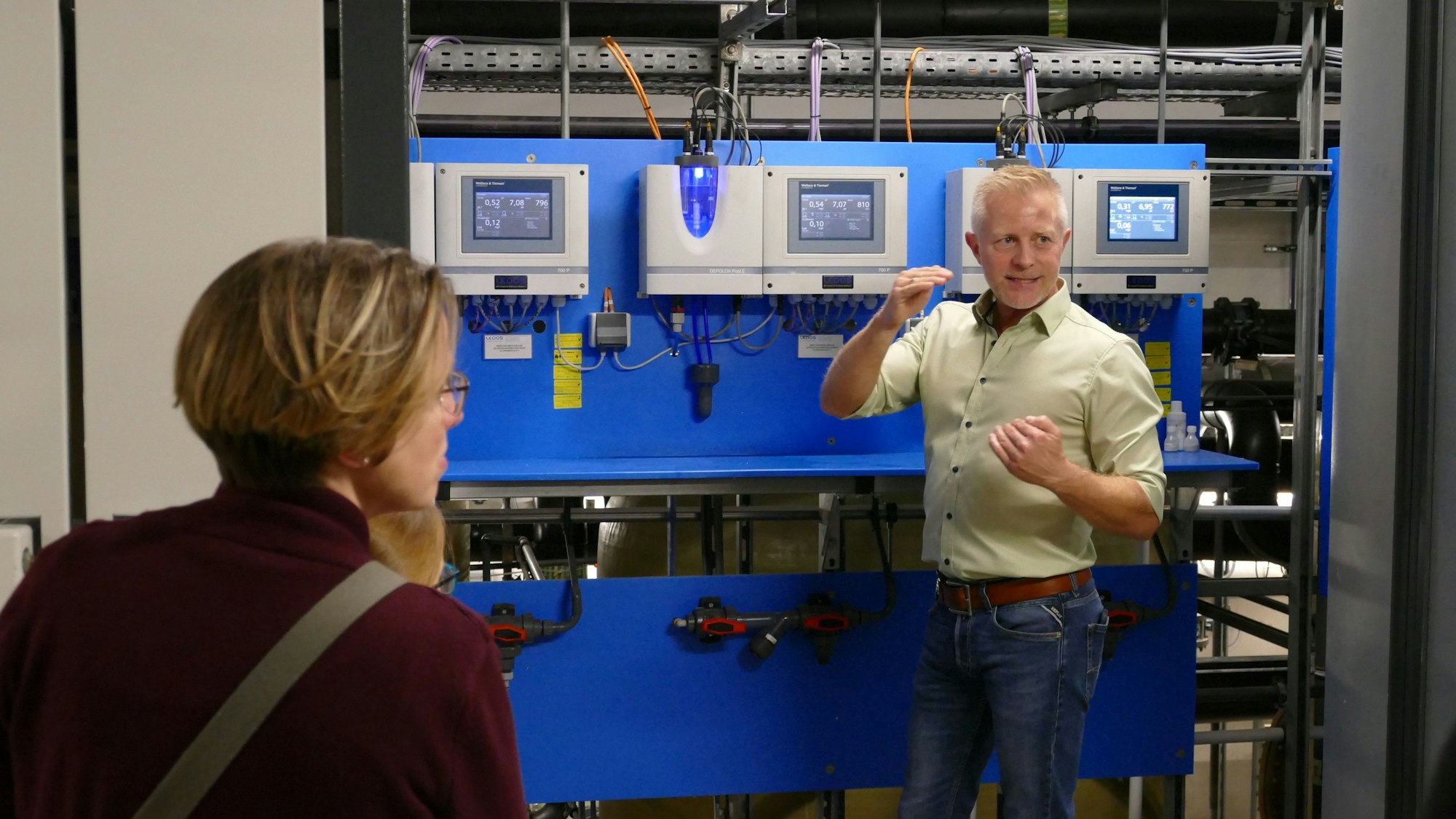 Ein Mann in einem beigefarbenen Hemd steht vor einer blauen Wand mit verschiedenen Digitalabzeigen.