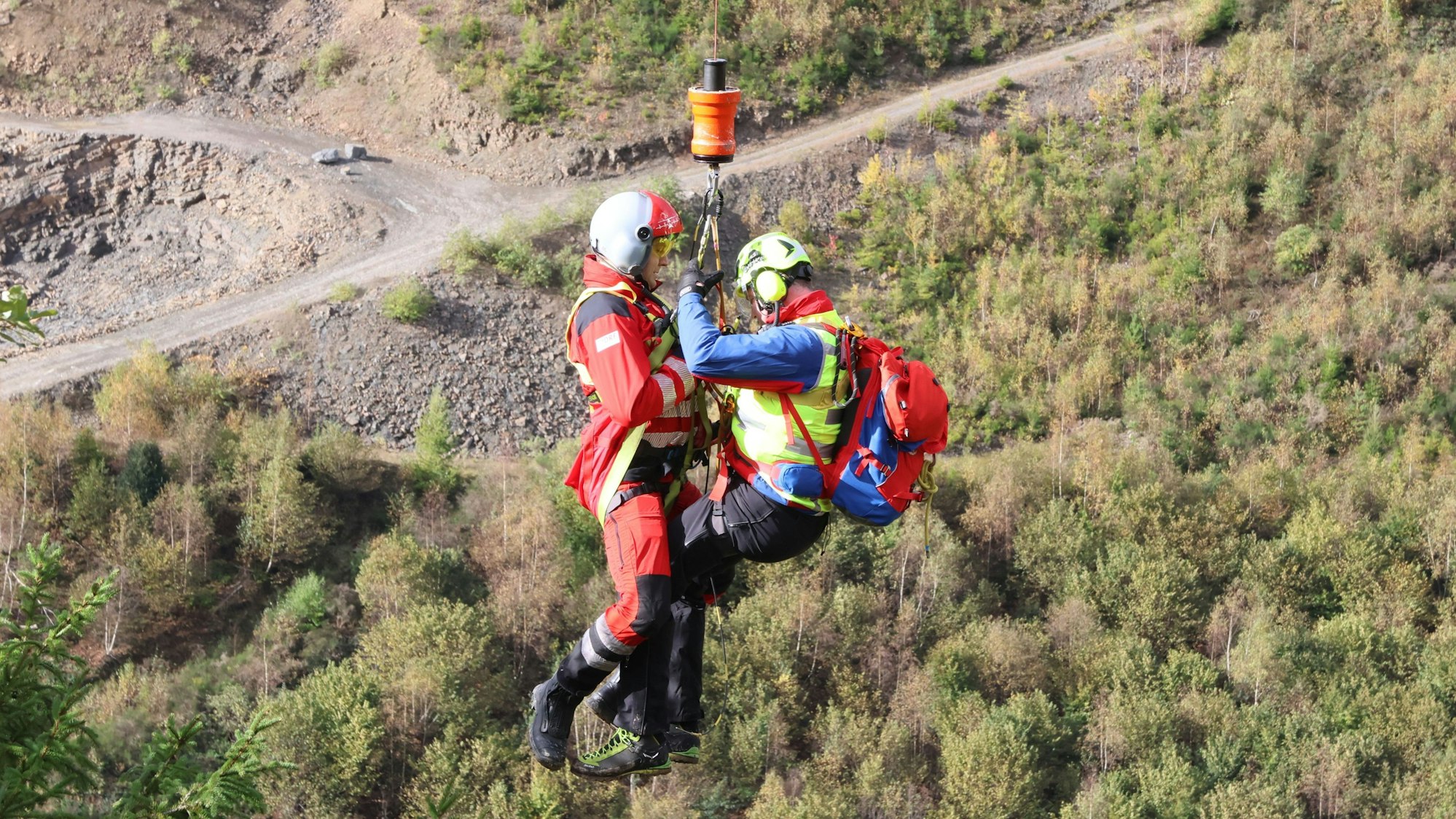 Zu sehen ist eine Übung der Bergwacht, bei der zwei Menschen von einer Hubschrauberwinde nach oben gezogen werden.