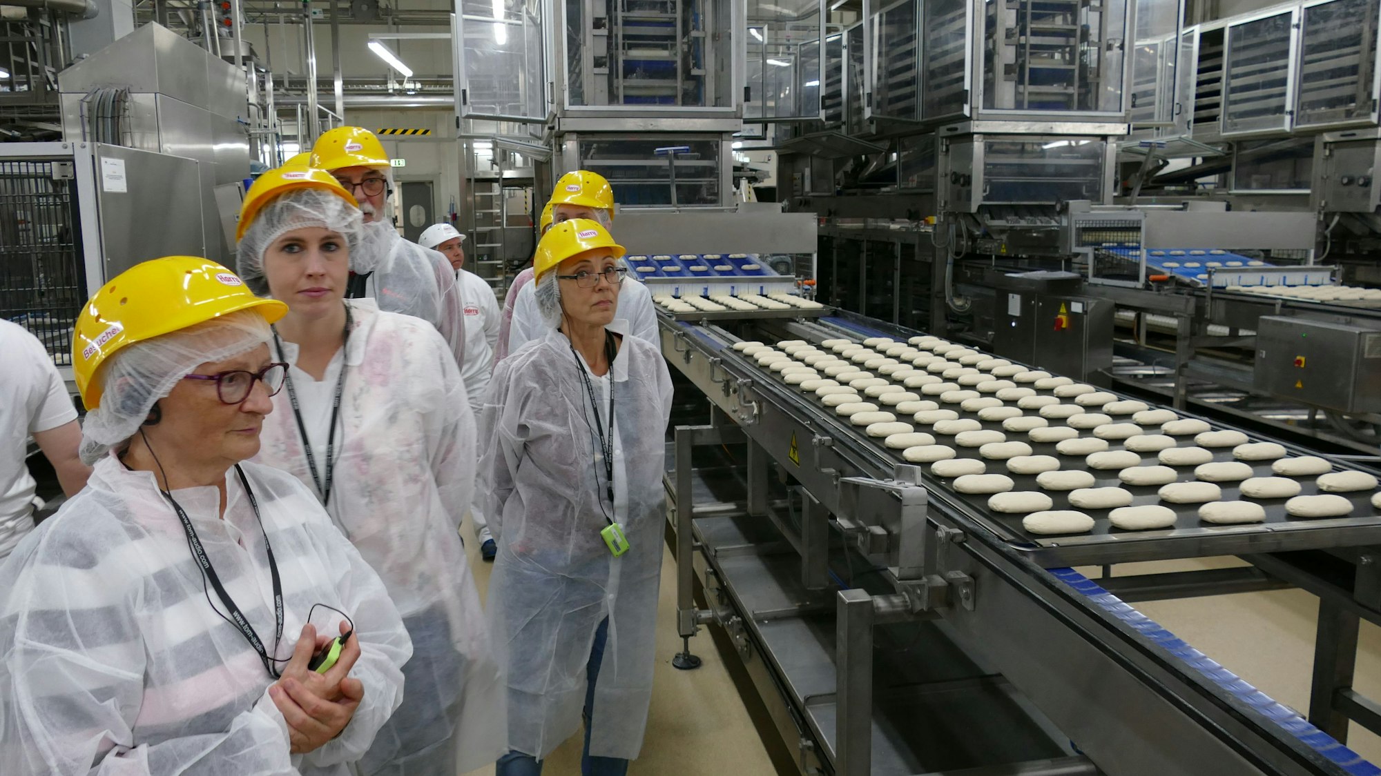 Menschen in weißen Schutzanzügen und mit gelben Sicherheitshelmen in einer Backfabrik.