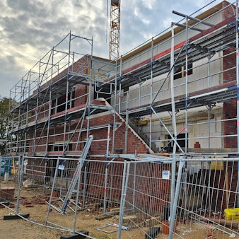 Ein Baugerüst umgibt ein großes, noch nicht fertiggestelltes Gebäude, das von einem Bauzaun geschützt ist.