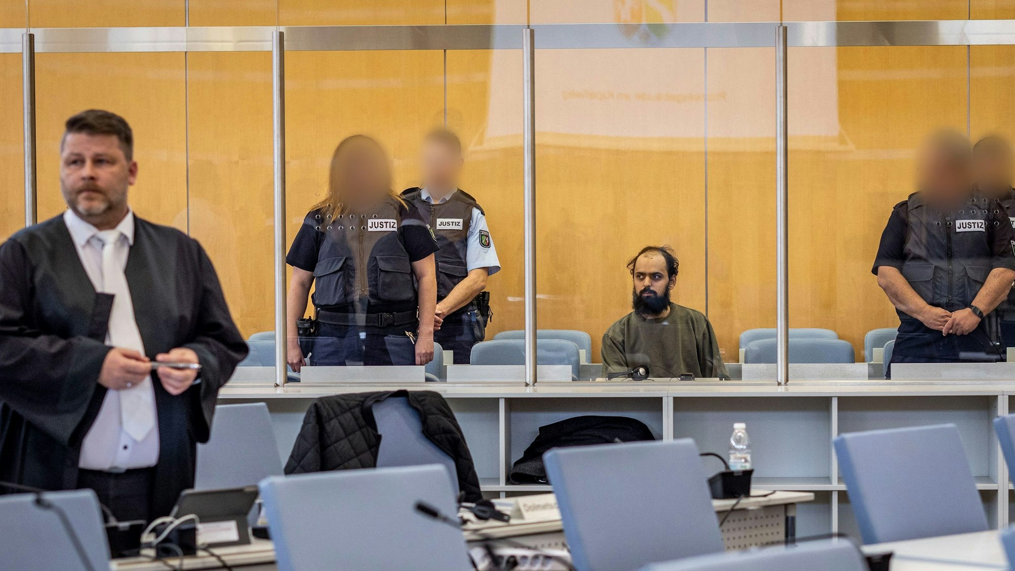 23.10.2023, Nordrhein-Westfalen, Düsseldorf: Der Angeklagte Maan D. (M) wird von Justizbeamten bewacht, er sitzt hinter einer Sicherheitsscheibe auf einem Stuhl. Links steht sein Anwalt Christian Grotenhöfer. ACHTUNG: Justizbeamte wurden auf Anweisung des Gerichts gepixelt +++ dpa-Bildfunk +++