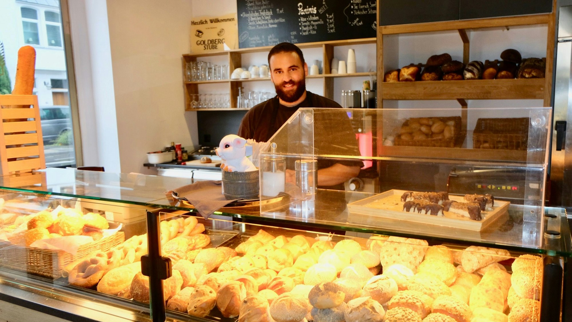 Ein Mann mit dunklem Haar und Bart steht hinter einer Bäcker-Theke.