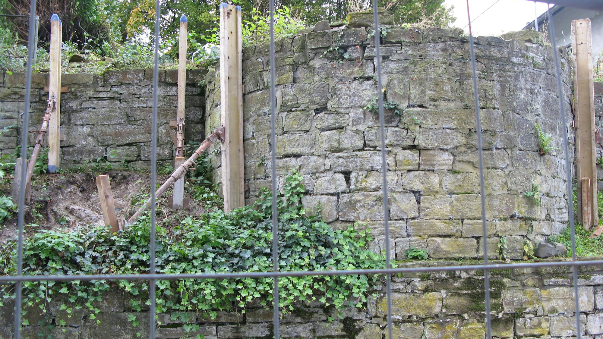 Im Vordergrund steht ein Bauzaun. Dahinter ist die halbrunde Apsis der zerstörten Synagoge zu sehen. Die Bruchsteinmauern sind durch Stützen gesichert.