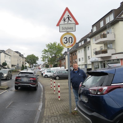 Ein Mann steht an einem Fußgängerüberweg. Autos fahren vorbei und parken am Straßenrand.
