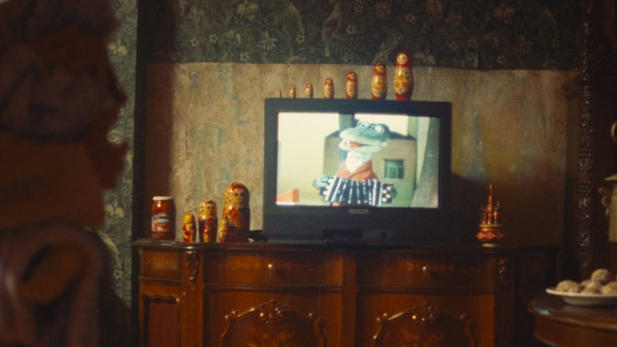 Das Krokodil Gena ist auf einem Röhrenfernseher zu sehen, auf dem und neben dem Matrjoschkas stehen.