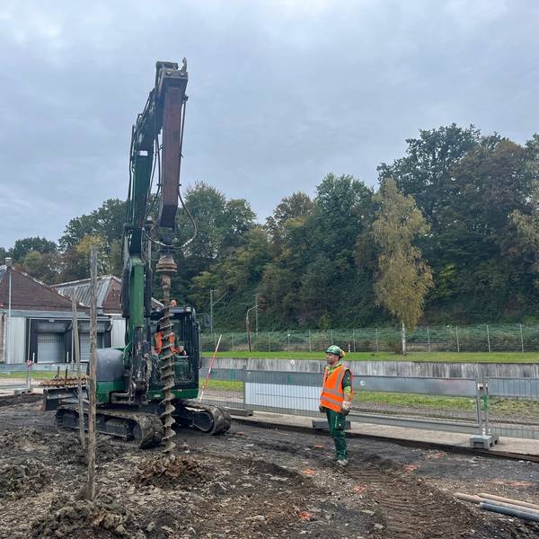 Ein Bagger vor Werkshallen mit einem Sheddach. Seitlich steht ein Arbeiter in roter Sicherheitsweste.