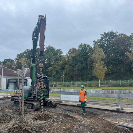 Ein Bagger vor Werkshallen mit einem Sheddach. Seitlich steht ein Arbeiter in roter Sicherheitsweste.