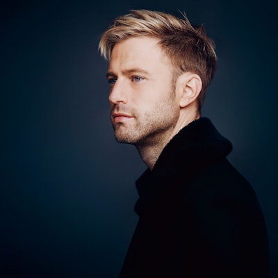 Appl ist auf dem Bild fast im Profil zu sehen, der Hintergrund ist udnkelblau. Er schaut verträumt , hat blondes Haar.