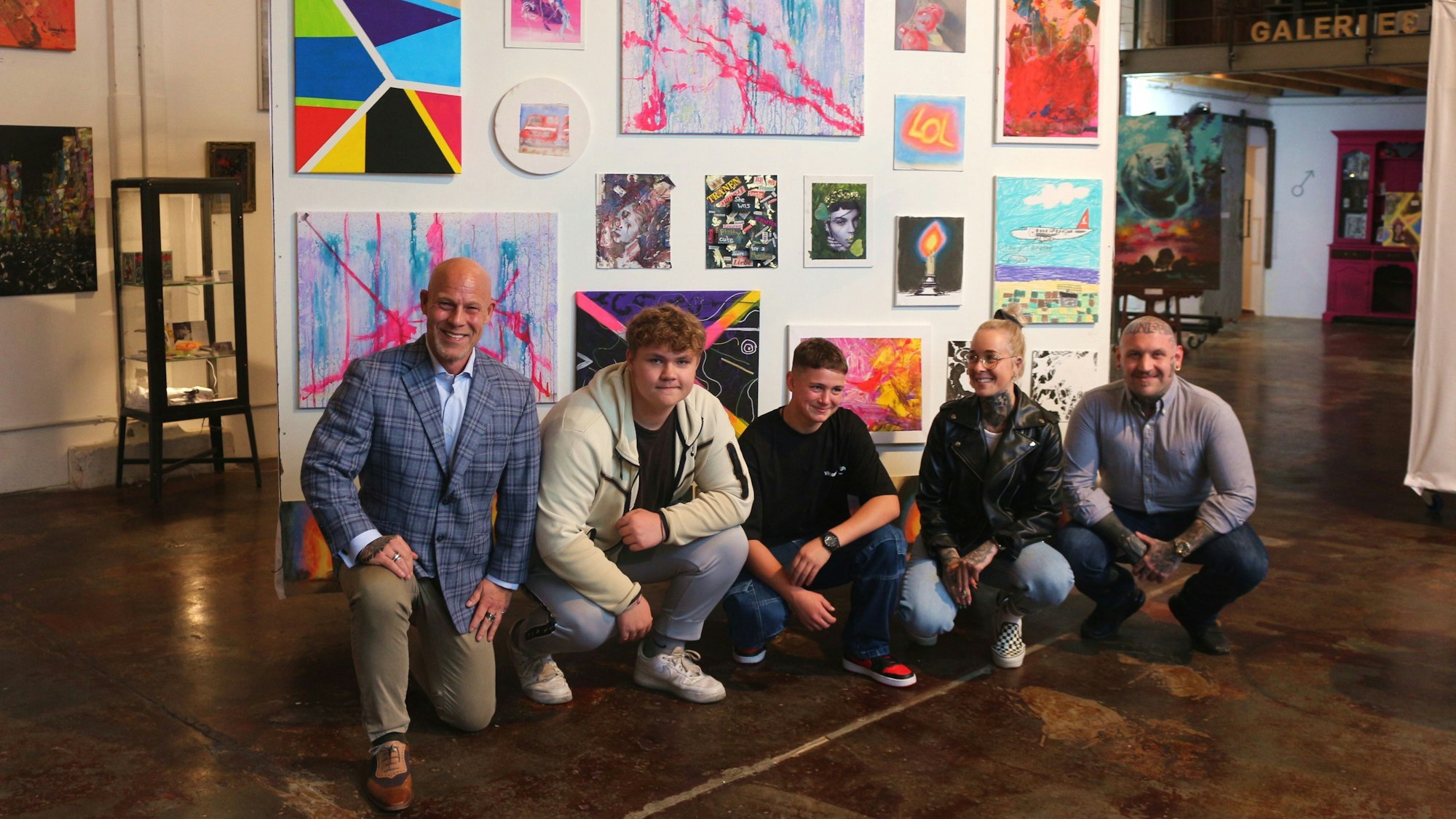 Zwei Männer, eine Frau und zwei Jugendliche knien vor einer Wand mit Gemälden.