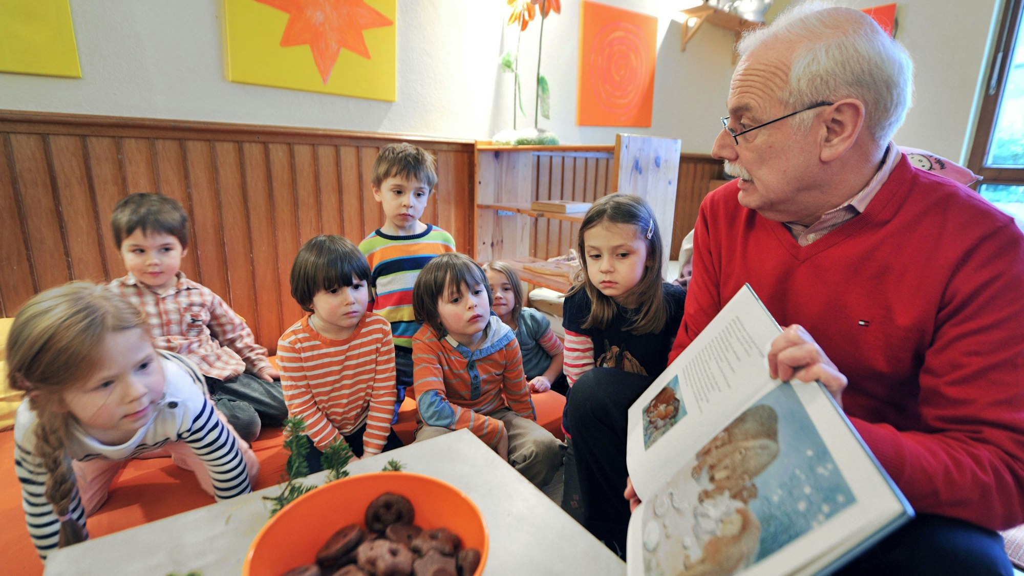 Am rechten Bildrand sitzt ein älterer Herr, der vor ihm sitzenden Kindergartenkindern Bilder eines Buches präsentiert, aus dem er vorgelesen hat. Die sieben Kinder schauen interessiert auf die Bilder.