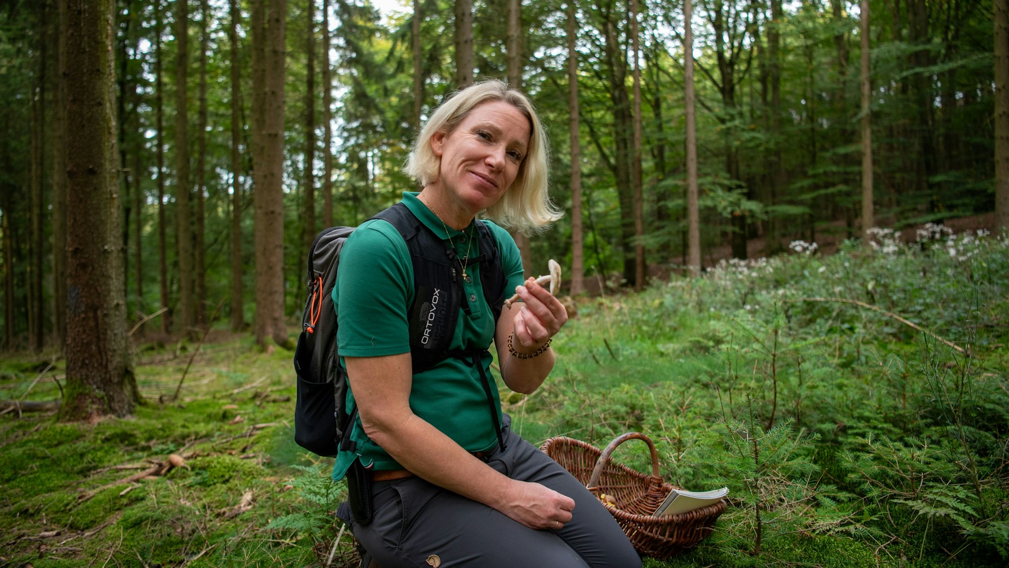 Christina Wojtowicz kniet auf dem Waldboden und hält einen Pilz in der Hand. Neben ihr steht ein Korb, in dem bereits gesammelte Pilze und ein Bestimmungsbuch liegen.