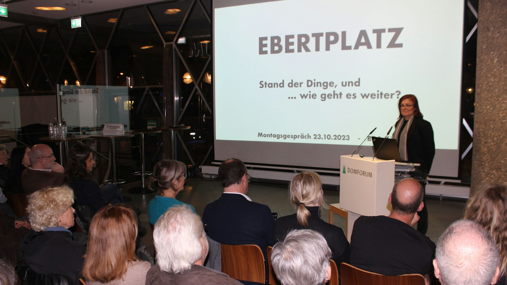 Eine Frau steht vor einer Leinwand auf einem Podium und vor ihr sitzen einige Menschen.