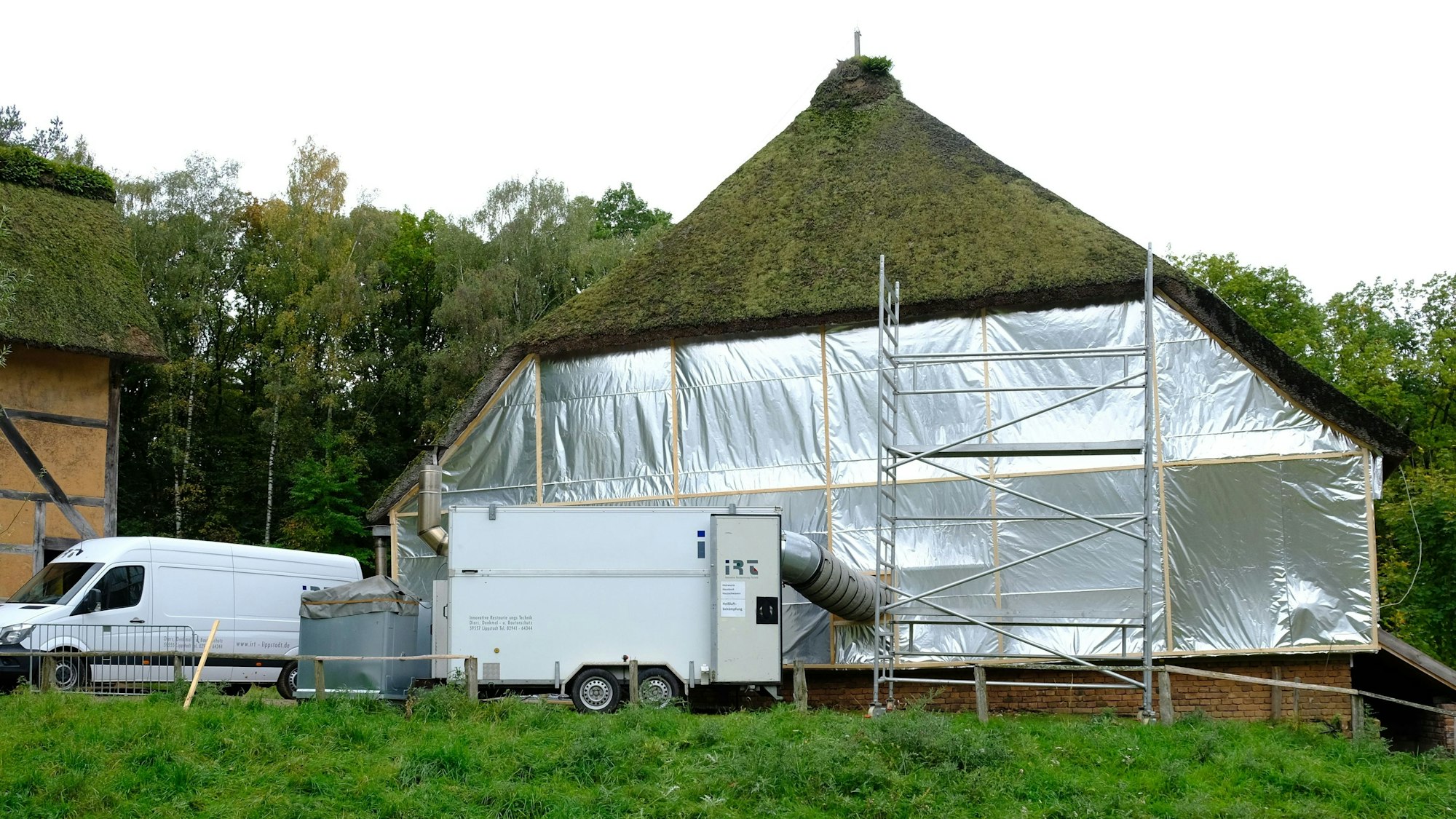 Bis auf das mit Reet gedeckte Dach ist ein großes Scheunengebäude im Freilichtmuseum Kommern in eine silbern glänzenden Folie eingepackt. Aus einem davor stehenden Anhänger führt ein voluminöser Schlauch ins Innere des Gebäudes.
