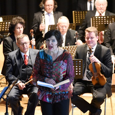 Eine Frau rezitiert vor einem Orchester stehend.