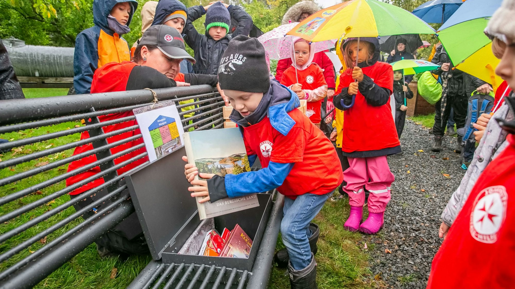 Kinder in Regekleidung stehen rund um eine Sitzbank im Freilichtmuseum. Ein Kind zieht ein Bilderbuch aus einer wetterfesten Truhe in der Bank.
