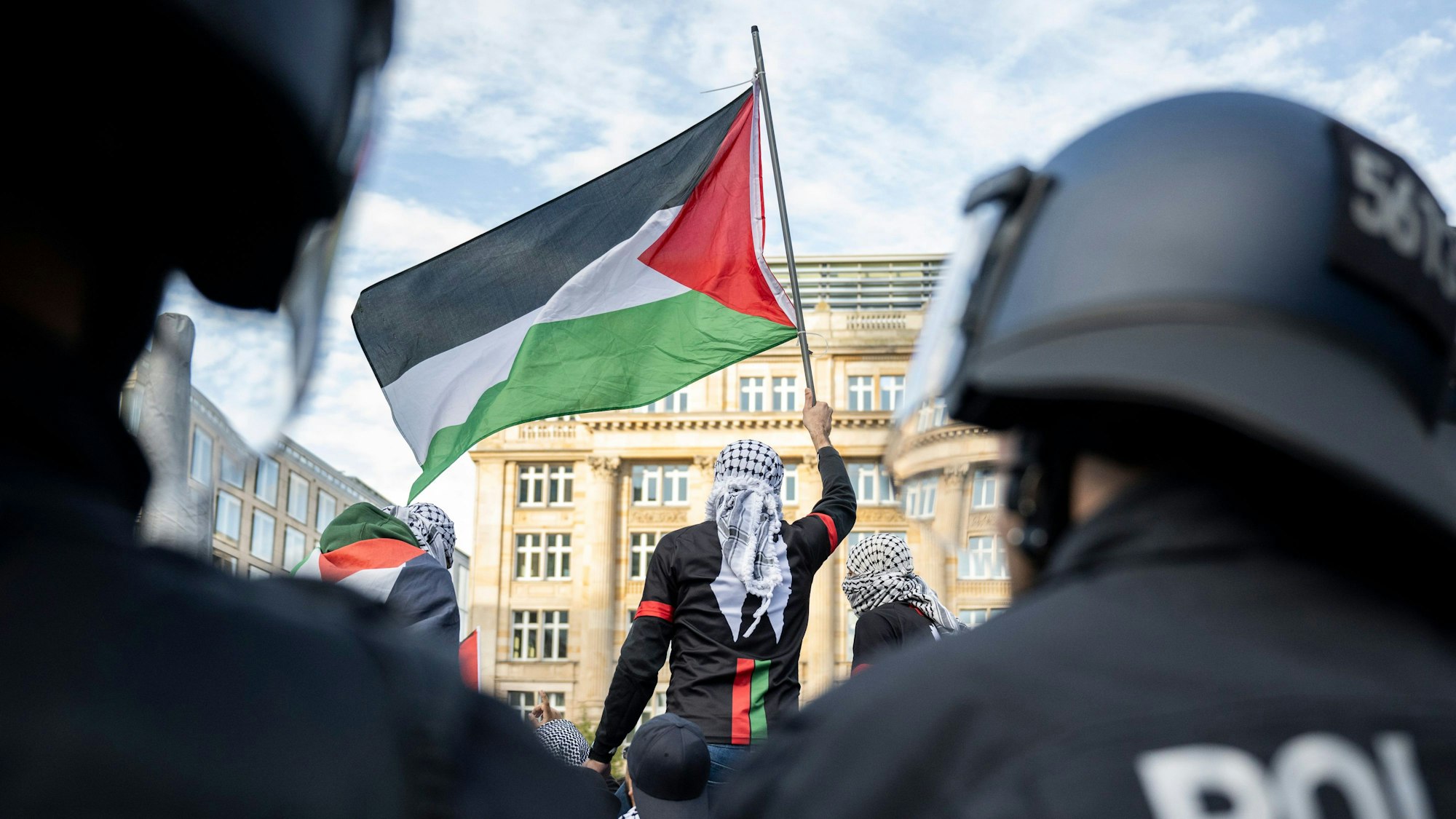 Ein Teilnehmer einer Pro-Palästina Demonstration schwenkt eine Palästina-Flagge, während Polizisten die Situation beobachten.