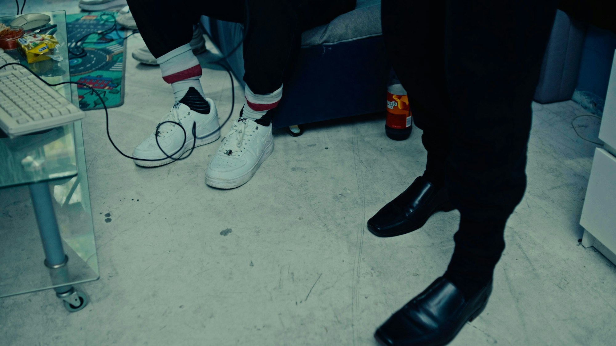 Das Bild zeigt die Schuhe von Equal und Liaze. Equal trägt weiße Sneaker und Tennissocken, in denen seine Hose steckt. Liazes Füße stecken in schwarzen Lederslippern, die er zur Jogginghose trägt.