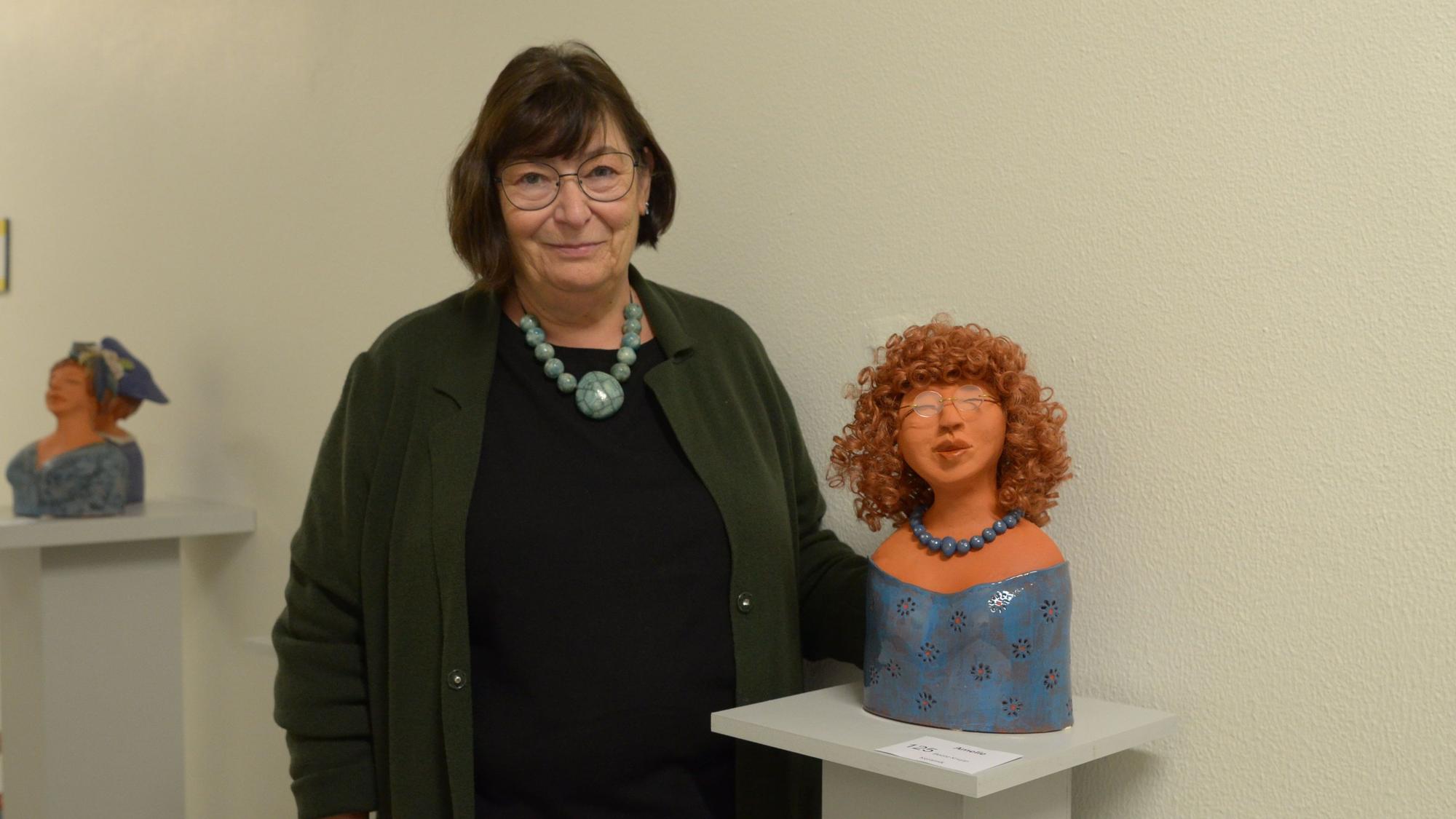 Die Künstlerin Beate Krupp steht neben einer ihrer Keramikskulpturen. Es handelt sich um eine Frauenfigur mit Perücke, Brille und Halskette.