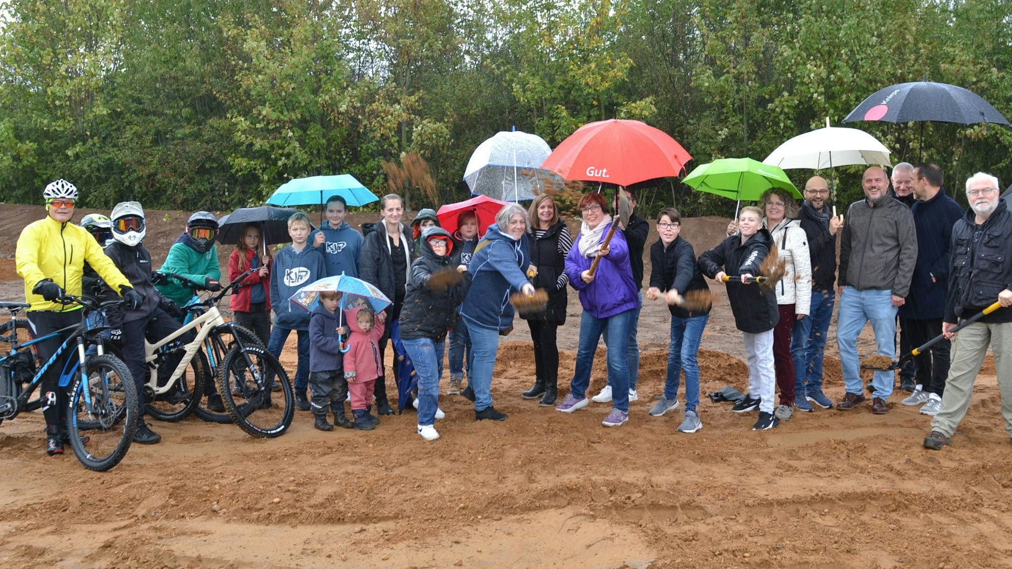 Eine größere Gruppe Menschen, Kinder und Erwachsene, steht mit Regenschirmen auf einem Bauplatz. Einige haben mit einem Spaten einen symbolischen Spatenstich vollzogen.