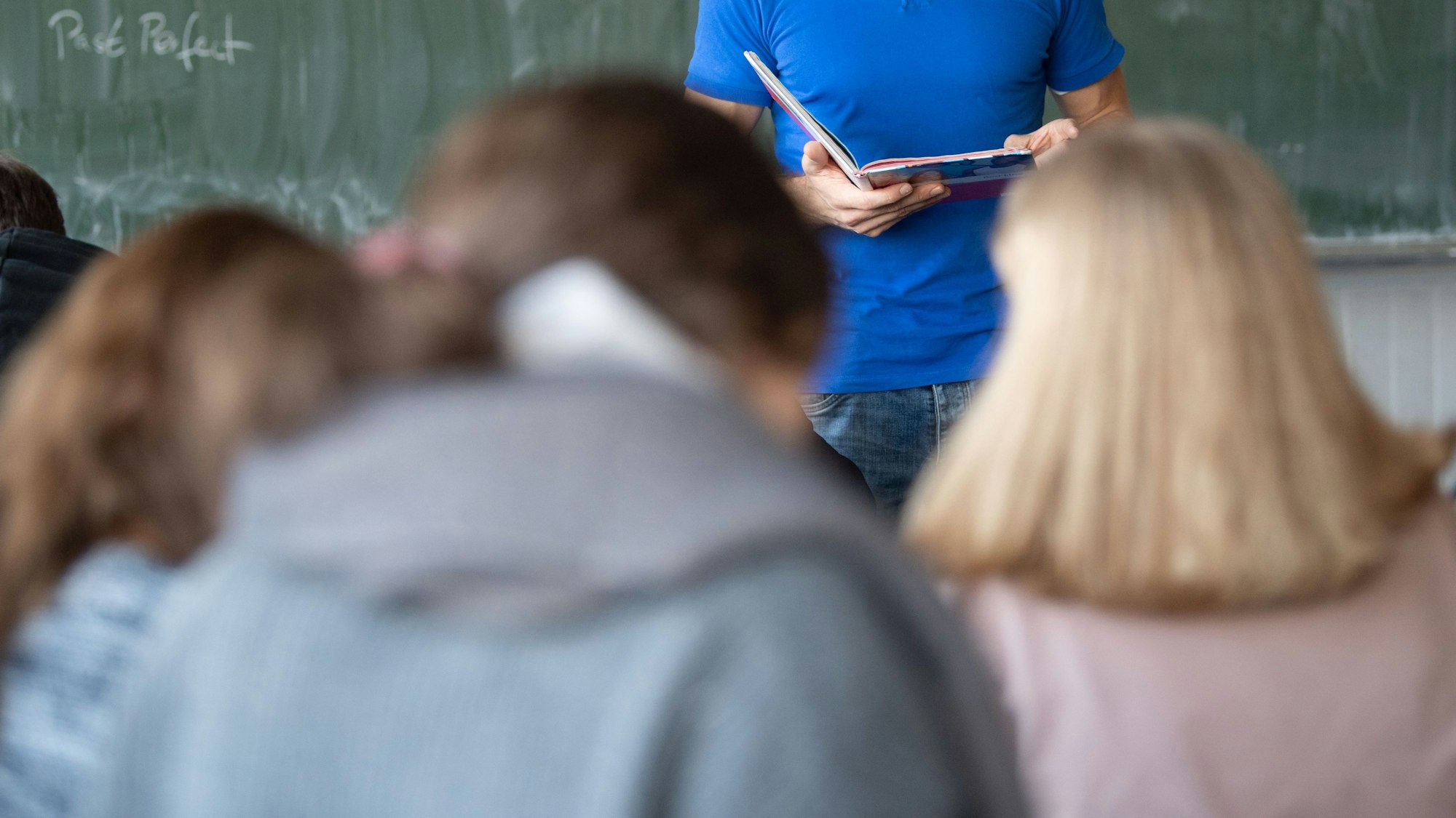 Symbolbild: Schülerinnen und Schüler in einem Klassenzimmer von hinten, im Hintergrund steht ein Lehrer vor einer Tafel.