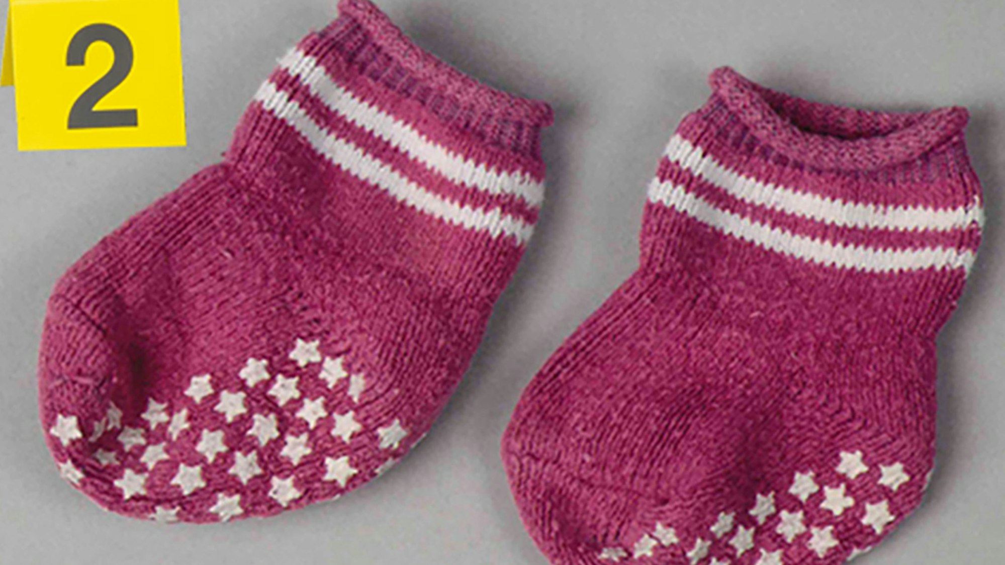 Zwei pinkfarbene Babysocken liegen auf einem Tisch. Danebensteht ein gelbes Schild mit der Nummer 2.