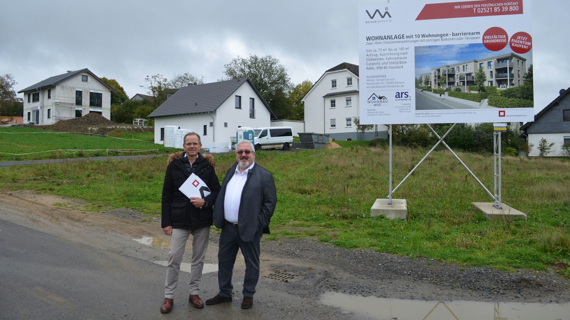 Zwei Männer stehen an einer Wiese, auf der ein großes Schild steht. Das weist darauf hin, dass dort ein Wohngebiet entstehen soll.