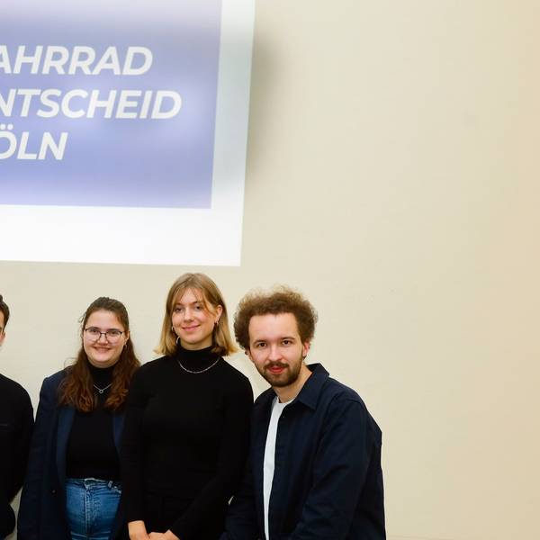 Zwei junge Männer und zwei Junge Frauen vor einem Plakat mit der Aufschrift „Fahrrad-Entscheid-Köln“.