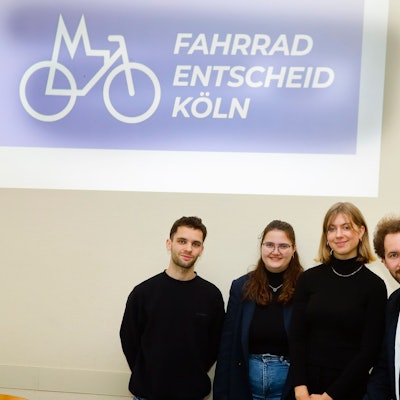 Zwei junge Männer und zwei Junge Frauen vor einem Plakat mit der Aufschrift „Fahrrad-Entscheid-Köln“.