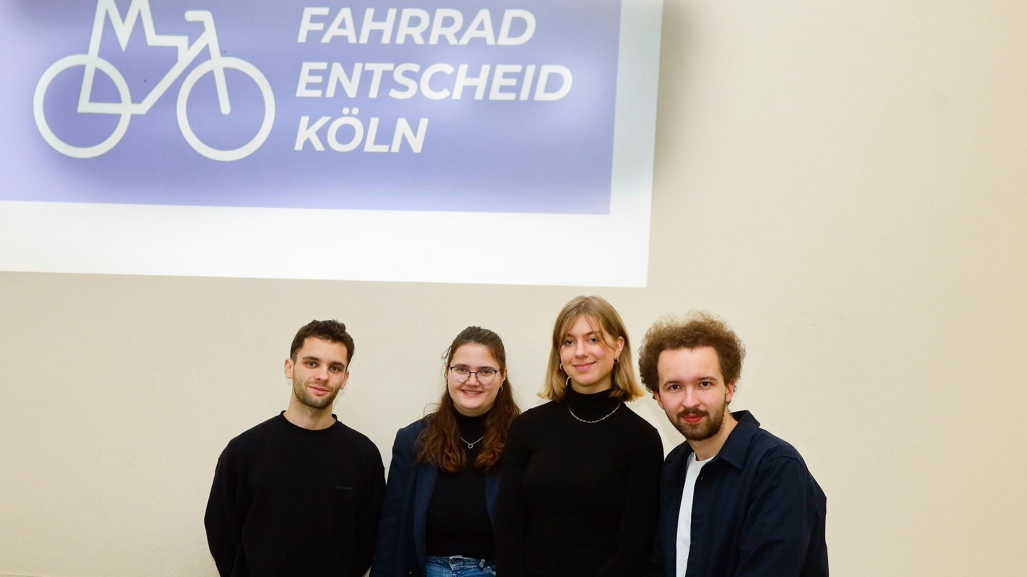 Zwei junge Männer und zwei Junge Frauen vor einem Plakat mit der Aufschrift „Fahrrad-Entscheid-Köln“.