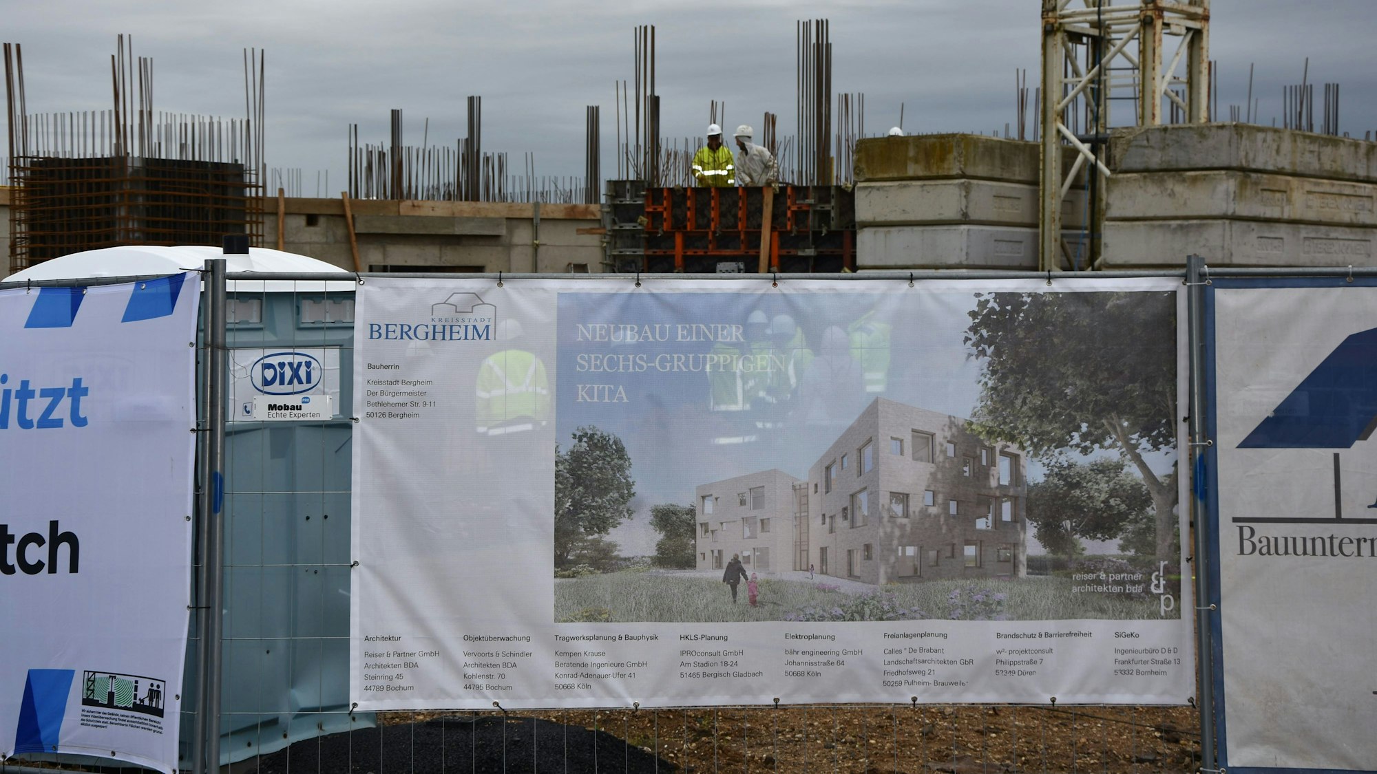 Auf dem Foto sind Bauarbeiter im Rohbau zu sehen. Davor ist das Bauschild zu sehen.