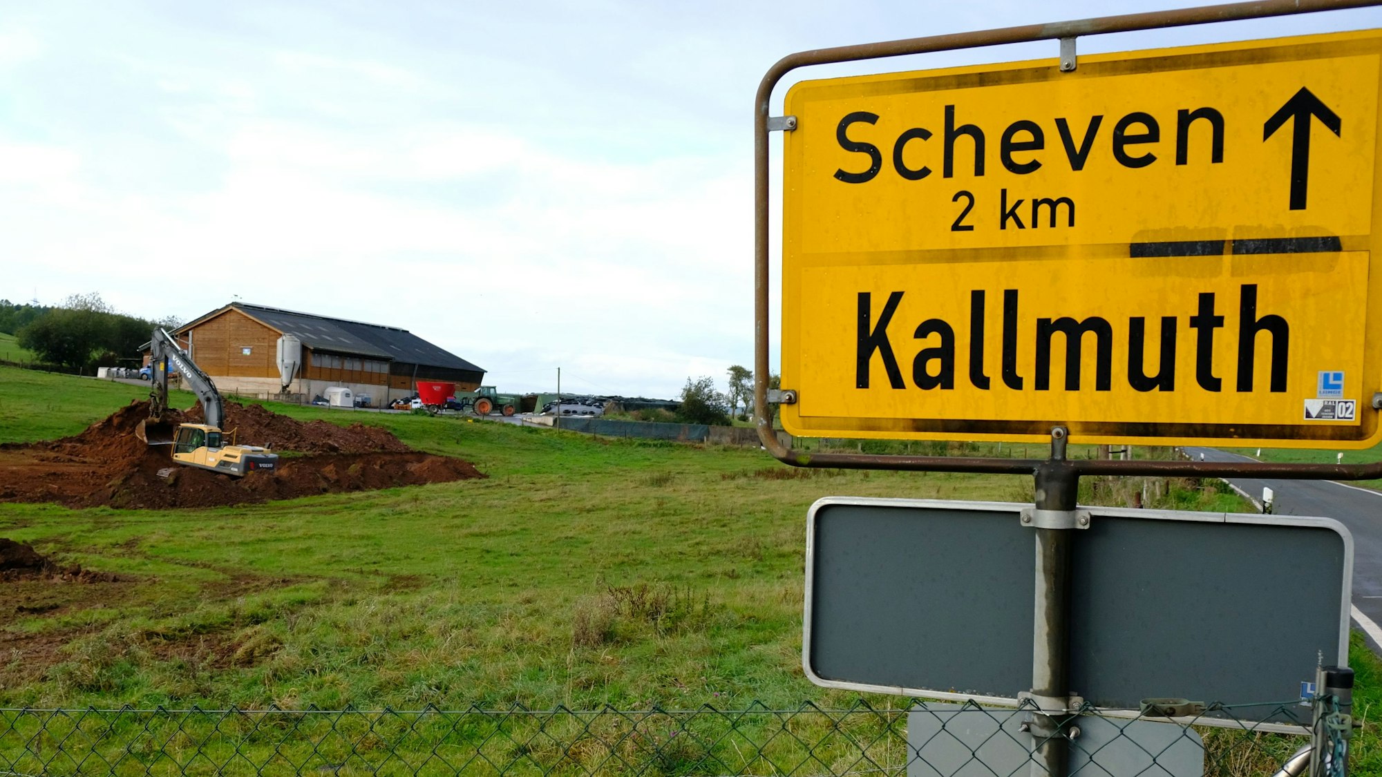 Im Hintergrund sind ein landwirtschaftliches Gebäude und ein Bagger bei Erdarbeiten auf einer Wiese zu sehen. Rechts das Ortsschild von Kallmuth.