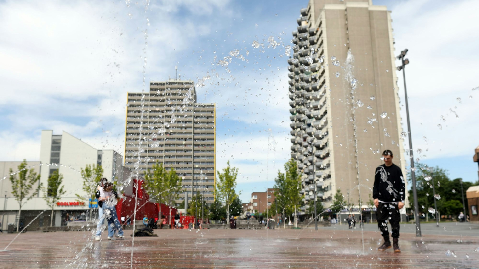 Aus dem Boden schießen Fontänen auf einem Platz, im Hintergrund sind Hochhäuser zu sehen.