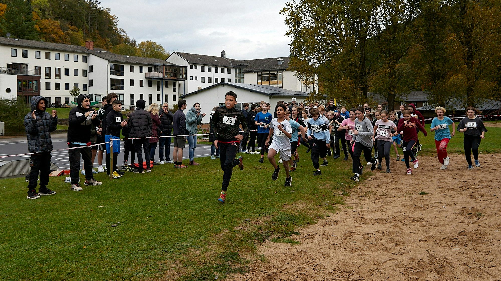 Zahlreiche junge Läufer starten zu einem Crosslauf. Hinter einem Flatterband stehen Jugendliche und Erwachsene und feuern sie an.