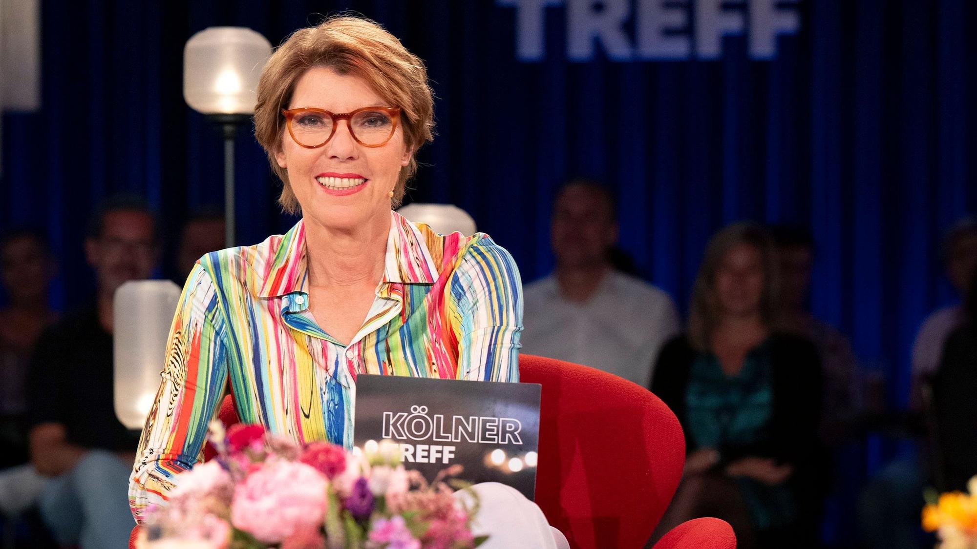 Moderatorin Bettina Böttinger sitzt im Studio des „Kölner Treff“. Sie trägt eine bunt gemusterte Bluse und blickt lächelnd in die Kamera.