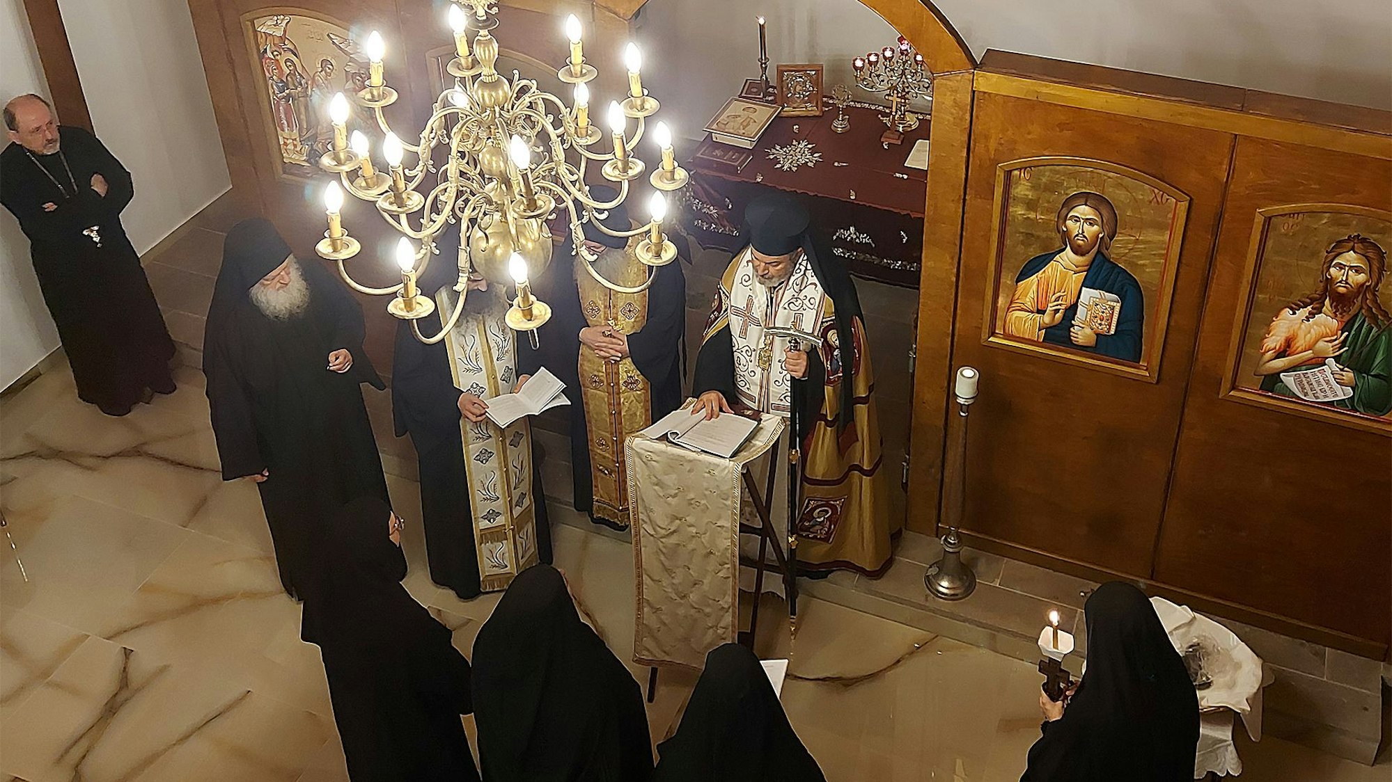 Orthodoxe Priester und Nonnen stehen in einer Kapelle unter einem Kronleuchter. An den holzvertäfelten Wänden sind sakrale Gemälde zu sehen.