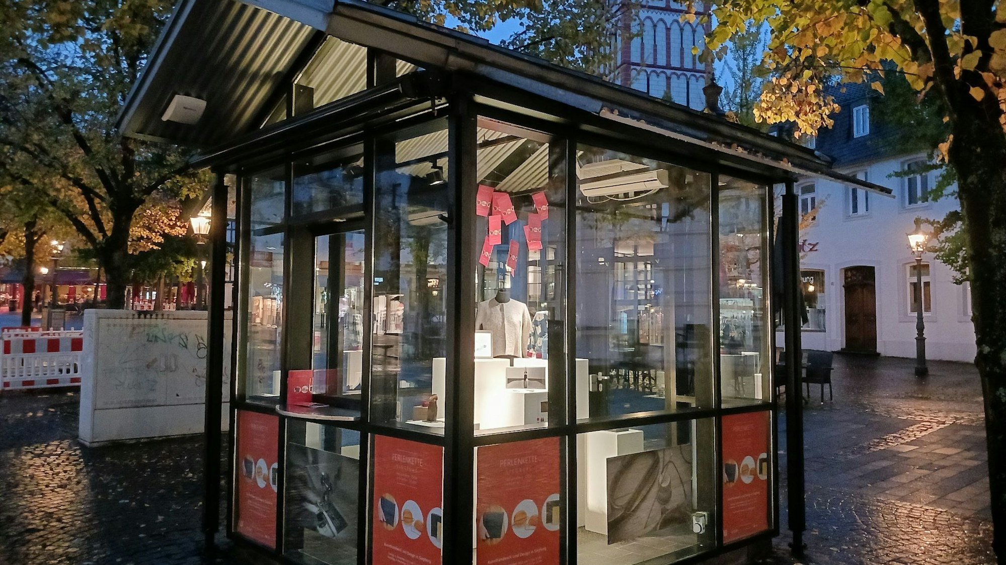 Der hellerleuchtete Kiosk auf dem Siegburger Markt, im Inneren Werbung für eine große Aktion mit Kunsthandwerk und Designartikeln