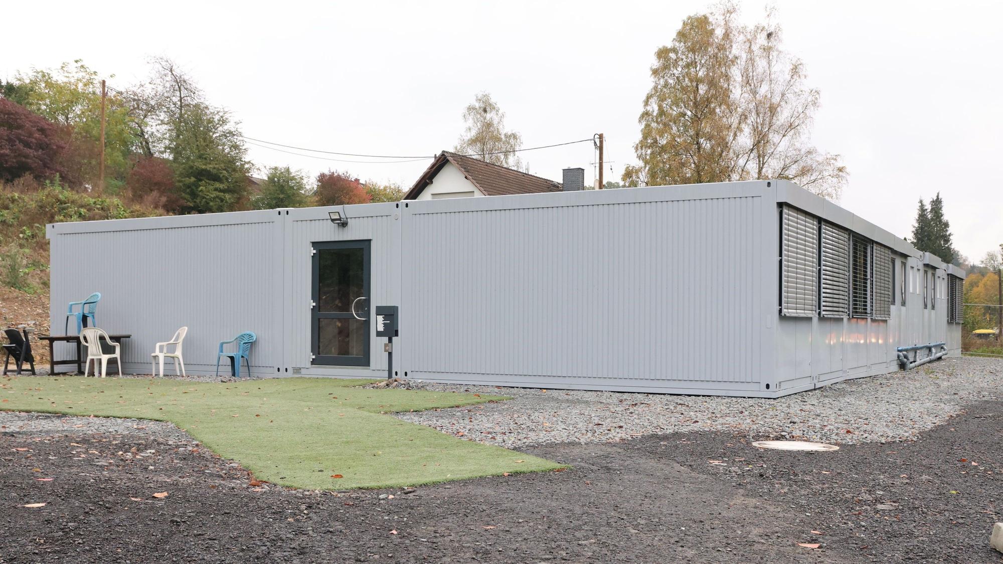 Vor einer Container-Unterkunft stehen leere Plastikstühle auf Kunstrasenteppichen.