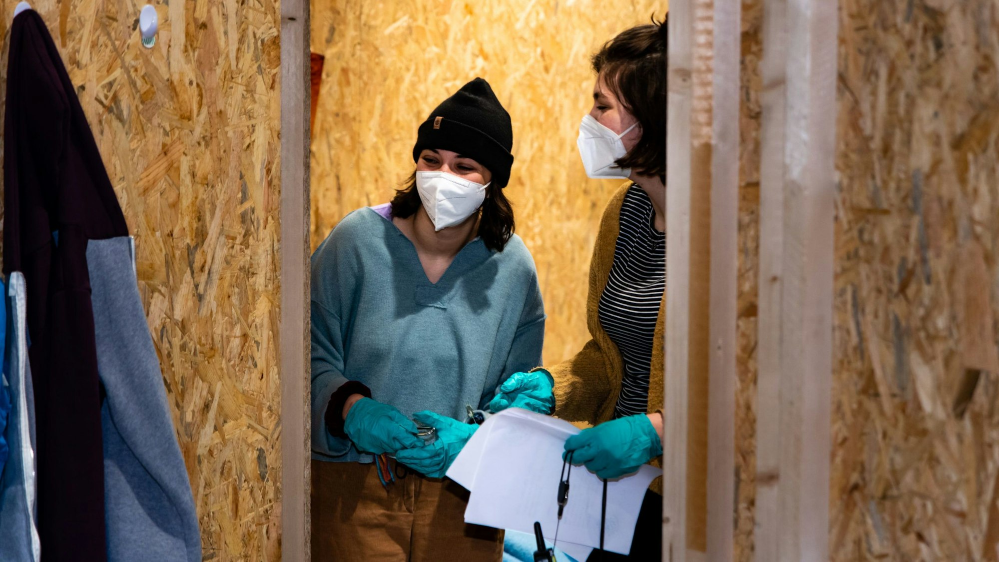 Zwei junge Frauen mit Masken und Einmalhandschuhen halten Papierblätter in der Hand und laufen durch den Escape-Room.