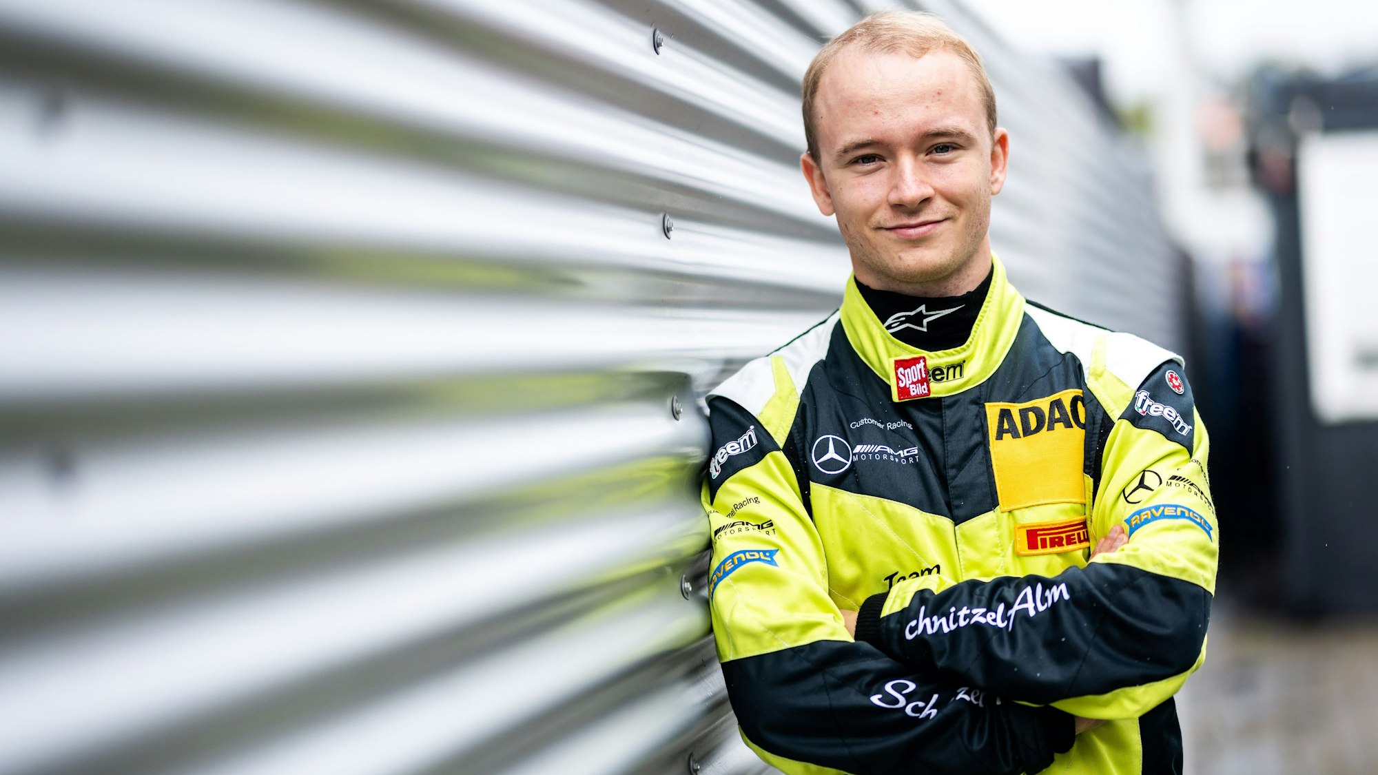 Moritz Wiskirchen lehnt in einem gelb-schwarzen Rennanzug an einer Wand aus Wellblech.
