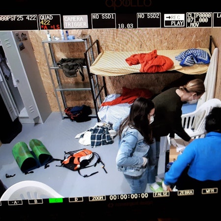 Drei Menschen suchen auf einem Tisch in einem kleinen Raum nach Unterlagen. Um sie herum liegen Kleidungsstücke, ein Rucksack und eine Isomatte auf dem Boden. In der Ecke des Raums steht ein Etagenbett aus Metall.